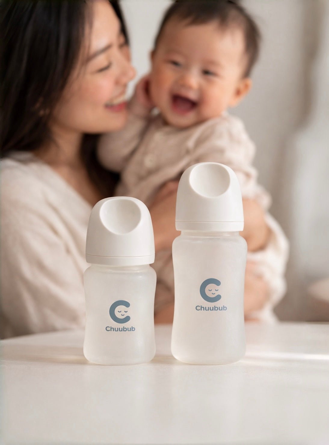 A mother holding her laughing baby beside two Chuubub SiliGlass milk bottles